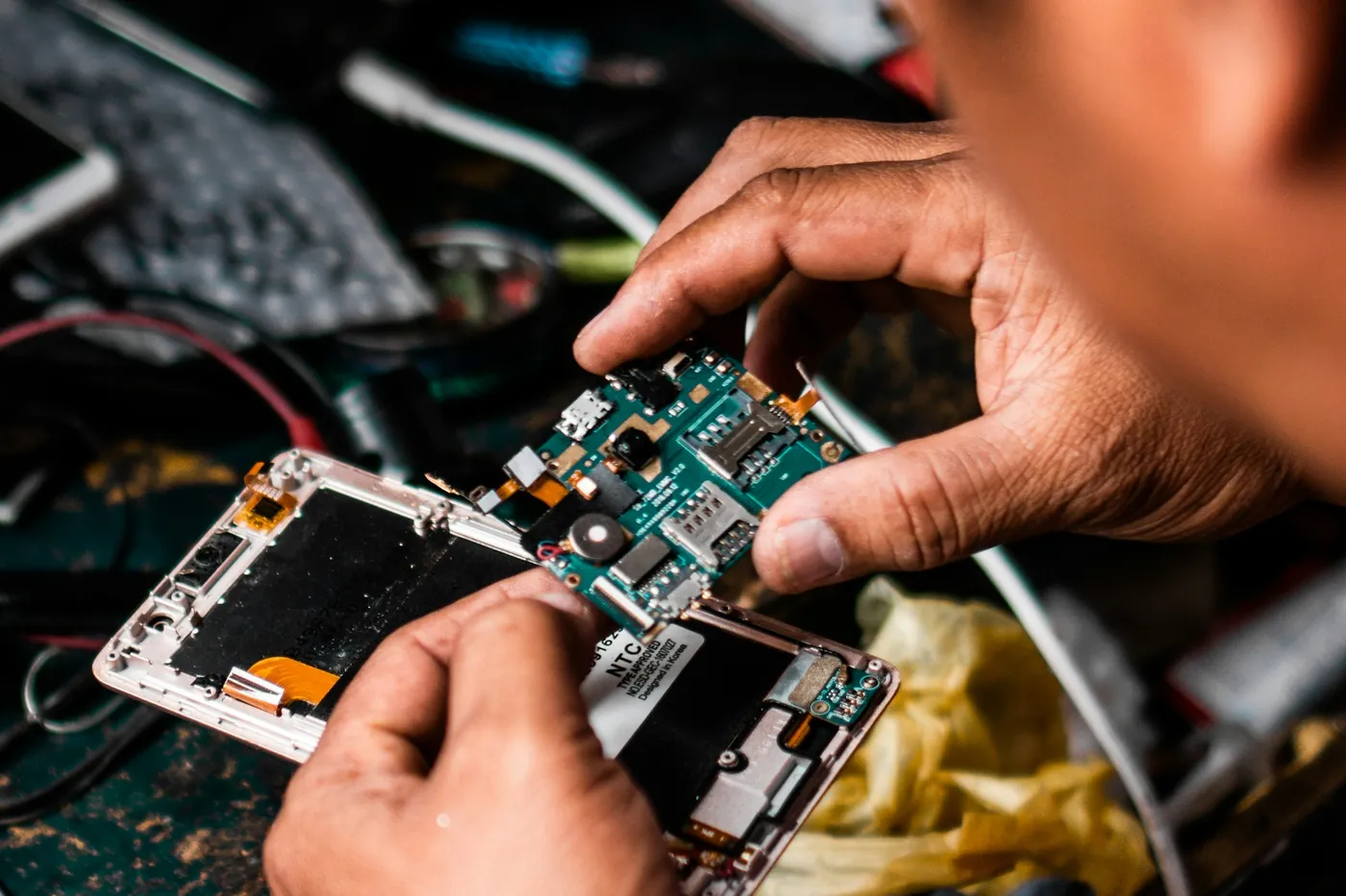 Mãos segurando a placa de um celular desmontado sobre a bancada, ao lado da carcaça com a tela